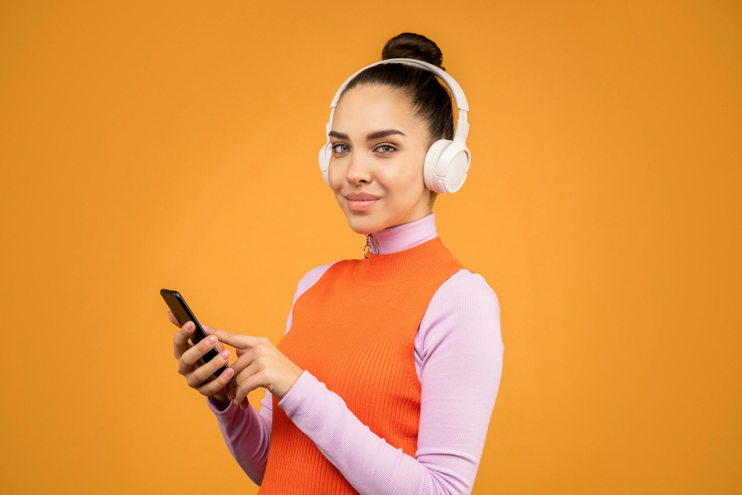 Portrait of a young woman with headphones and smartphone against a vibrant orange background.